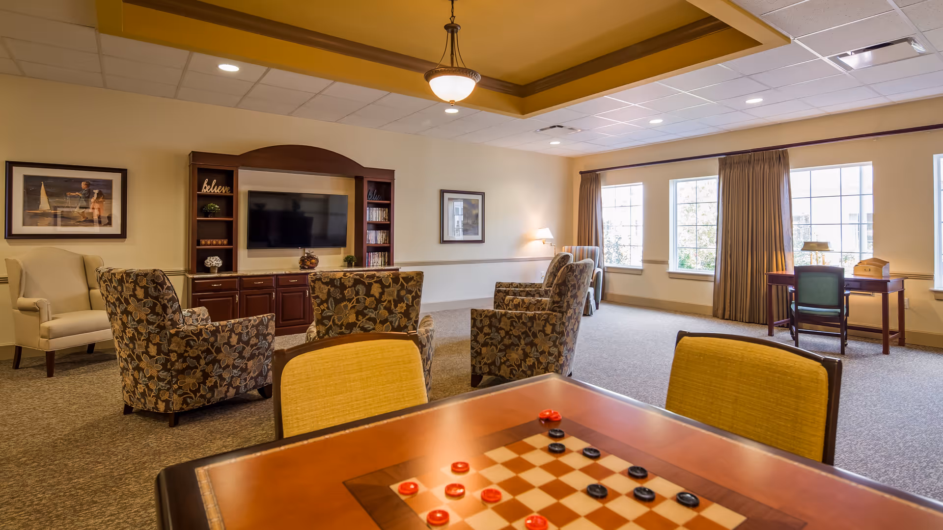 A spacious living room area with a checkers game table in the foreground, several upholstered armchairs arranged facing a wall-mounted flat screen TV, a wooden entertainment center, framed artwork on the walls, large windows with curtains letting in natural light, and a small desk with a chair near the windows.