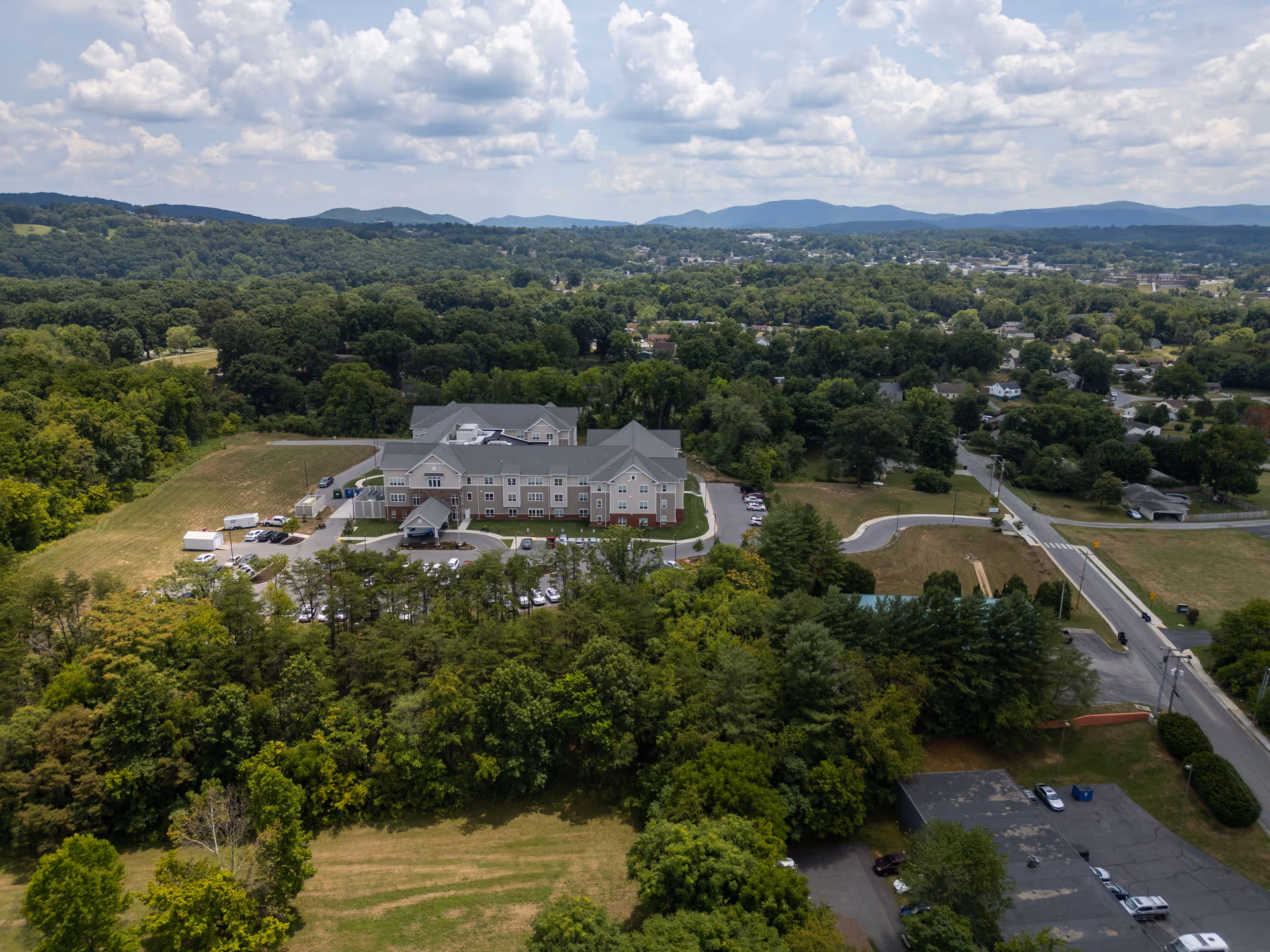 Aerial view of a multi-story nursing facility building surrounded by trees, parking lots, roads, and distant hills under a partly cloudy sky.