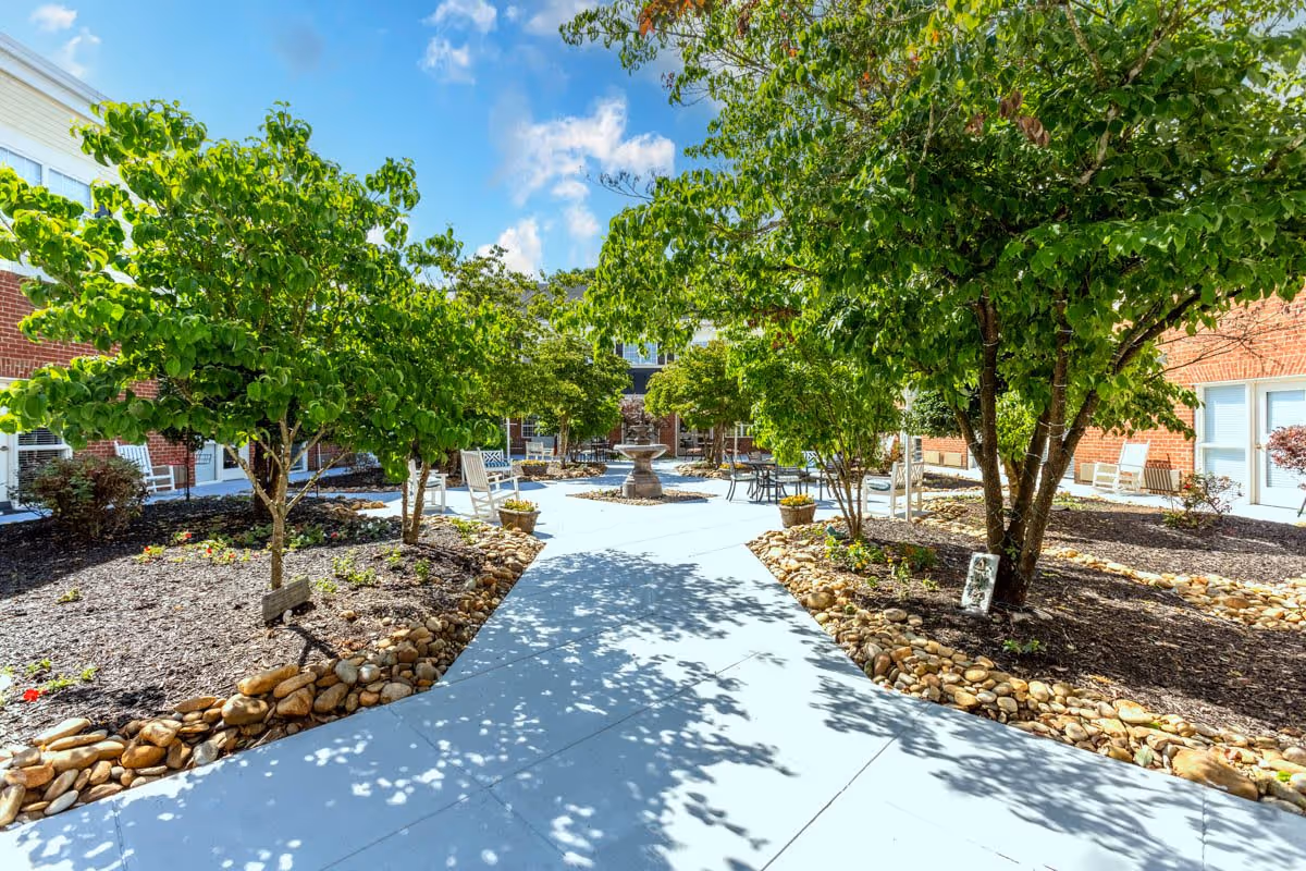 Outdoor courtyard area at StoryPoint Knoxville West with a concrete walkway, green leafy trees, a central water fountain, and seating areas including white rocking chairs and tables with chairs. The courtyard is surrounded by red brick buildings under a blue sky with some clouds.