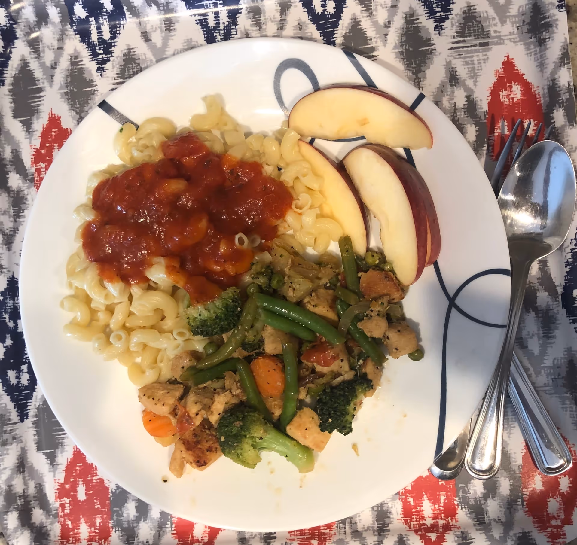 A white plate with a serving of elbow macaroni topped with red tomato sauce, a portion of mixed vegetables including broccoli, green beans, carrots, and pieces of chicken, and three slices of red apple. The plate is placed on a patterned tablecloth with red, gray, and blue designs. A spoon and fork are placed to the right of the plate.
