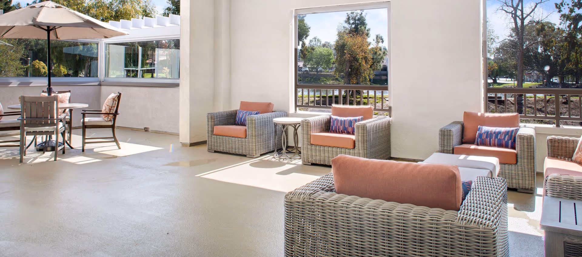 A bright and spacious outdoor patio area with wicker armchairs featuring orange cushions and patterned pillows, a round table with chairs under a large beige umbrella, and large windows showing trees and greenery outside.