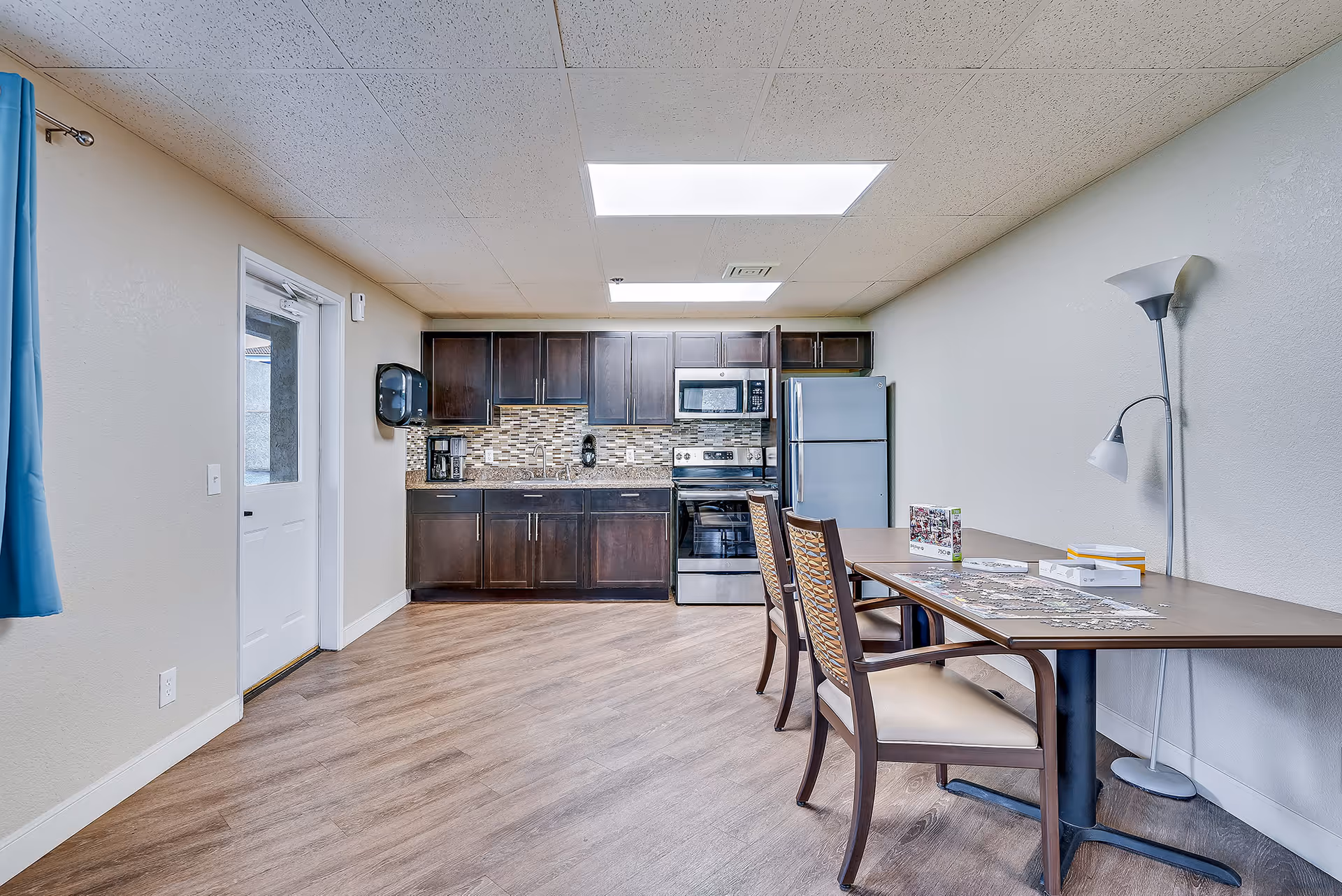 A bright kitchen area with dark wood cabinets, a stainless steel refrigerator, stove, and microwave. There is a granite countertop with a coffee maker and a sink. To the right, there is a table with two chairs and a floor lamp next to it. The floor is wood, and there is a door with a window on the left side of the room.
