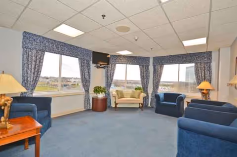 A spacious senior living lounge with blue seating, large windows, and patterned curtains.
