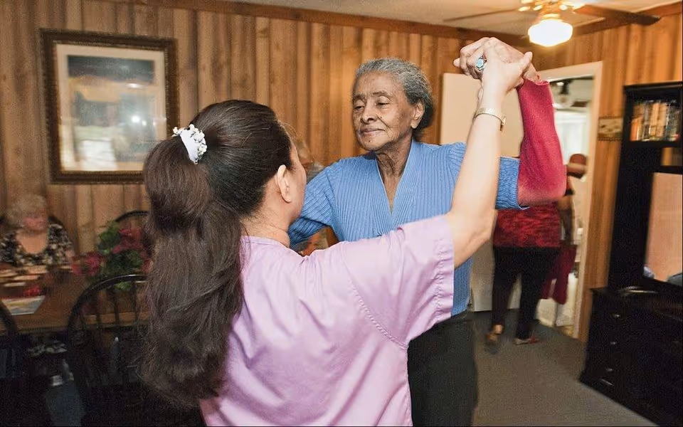 An elderly woman in a blue sweater is dancing with a caregiver wearing a pink uniform in a wood-paneled room. In the background, another elderly woman is seated at a table, and a person is walking away through a doorway. The room has a ceiling fan and framed artwork on the wall.