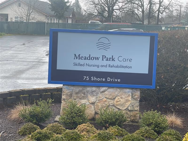 Outdoor sign for Meadow Park Care reading 'Skilled Nursing and Rehabilitation' and '75 Shore Drive' mounted on a stone base with shrubs and a parking lot behind it.