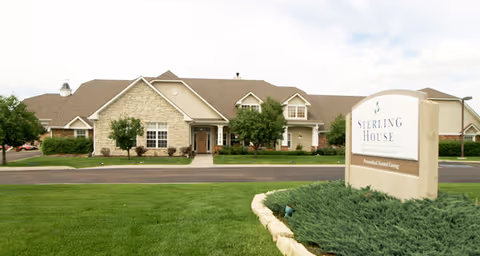 Exterior view of a single-story senior living facility building with beige stone and siding, a brown shingled roof, and several windows. There is a well-maintained lawn and landscaping in front, along with a large sign that reads 'Sterling House Senior Living Personal and Assisted Living.' The sky is partly cloudy.