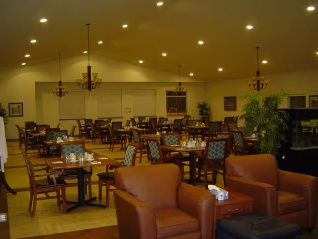 Spacious dining room with multiple set tables and chairs, chandeliers, and two armchairs in the foreground.