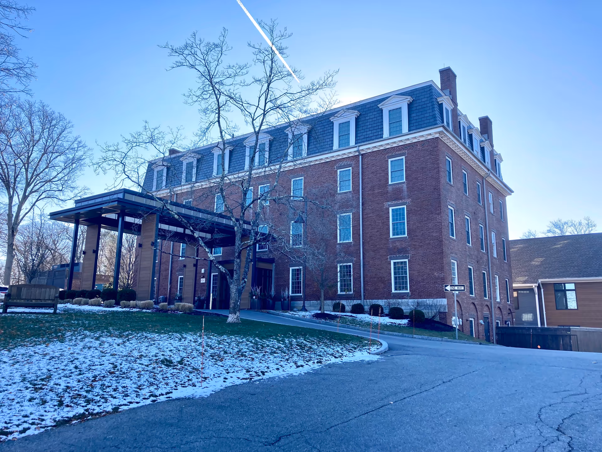 Exterior view of a large brick building with multiple windows and a covered entrance. There is a driveway leading up to the entrance, some leafless trees, a bench on the grass, and patches of snow on the ground under a clear blue sky.