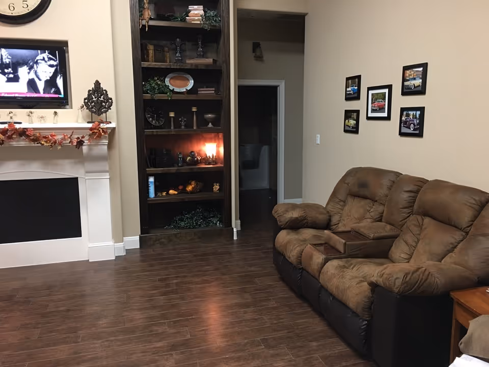 A cozy living room with a brown cushioned recliner sofa, a wooden floor, and a white fireplace decorated with autumn leaves. A TV is mounted above the fireplace showing a black and white image. There is a tall wooden bookshelf with various decorative items and a small light inside. On the wall above the sofa, there are five framed pictures of classic cars. A doorway leads to another room in the background.