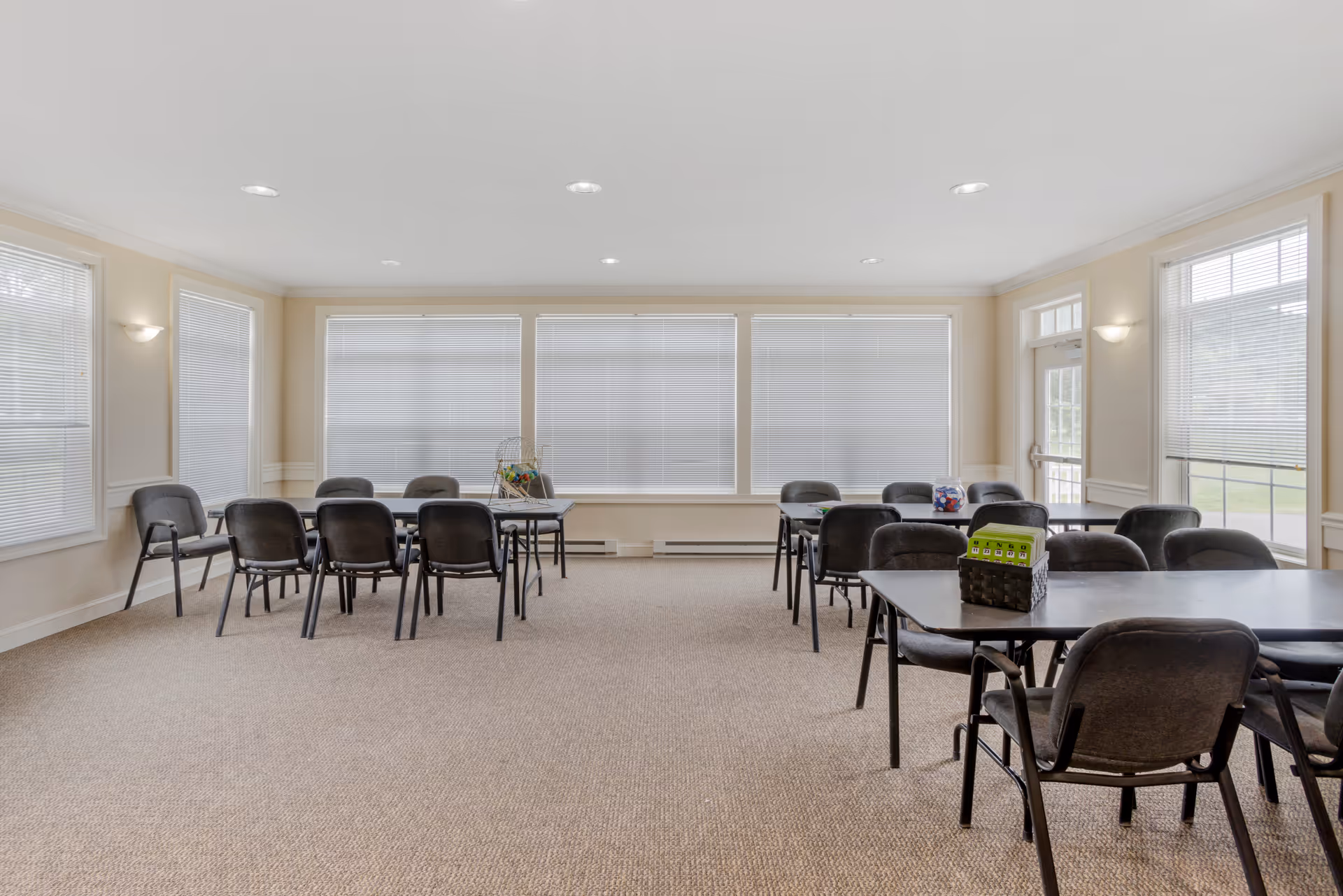 A bright room with beige walls and carpeted floor featuring several black tables and chairs arranged in groups. Large windows with white blinds cover the walls, allowing natural light to fill the room. On the tables, there are items such as a bingo cage and a container with bingo balls, suggesting the room is used for activities or gatherings.