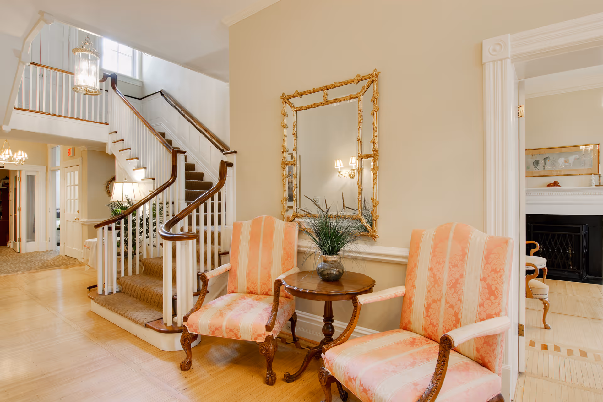 Interior view of a senior living facility showing a staircase with carpeted steps and wooden handrails. Two upholstered armchairs with pink and cream striped fabric flank a small wooden table with a potted plant. A large ornate gold-framed mirror hangs on the wall above the table. The area is well-lit with natural light from a window above the stairs and a hanging lantern-style light fixture. In the background, a hallway and a room with a fireplace and chairs are visible.