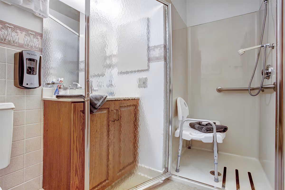 A bathroom with a wooden vanity and sink, a frosted glass shower door, and a walk-in shower equipped with a white shower chair and grab bar. There is a soap dispenser mounted on the tiled wall next to the sink.