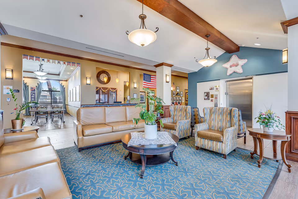 Bright senior living facility lobby with leather sofas, patterned armchairs, a central coffee table with a plant, and a dining area visible in the background.