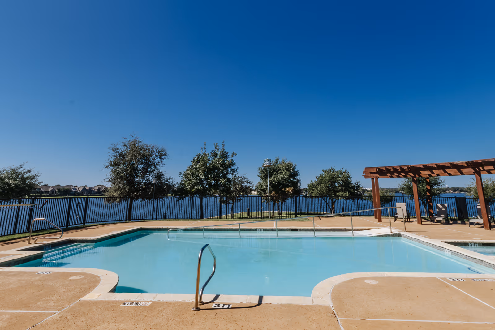 Outdoor swimming pool with clear blue water, surrounded by a concrete deck and metal handrails. In the background, there is a black metal fence, several trees, and a large body of water under a clear blue sky. To the right, there is a wooden pergola with chairs underneath.