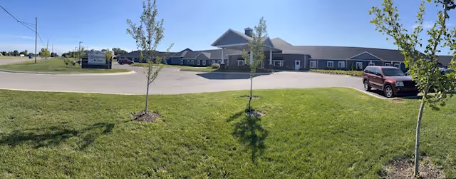 Wide panoramic view of the front exterior of Hampton Manor of Trenton Premier Assisted Living Facility, showing a single-story building with a peaked roof, a parking lot with a red SUV, young trees planted on a grassy lawn, and a clear blue sky.