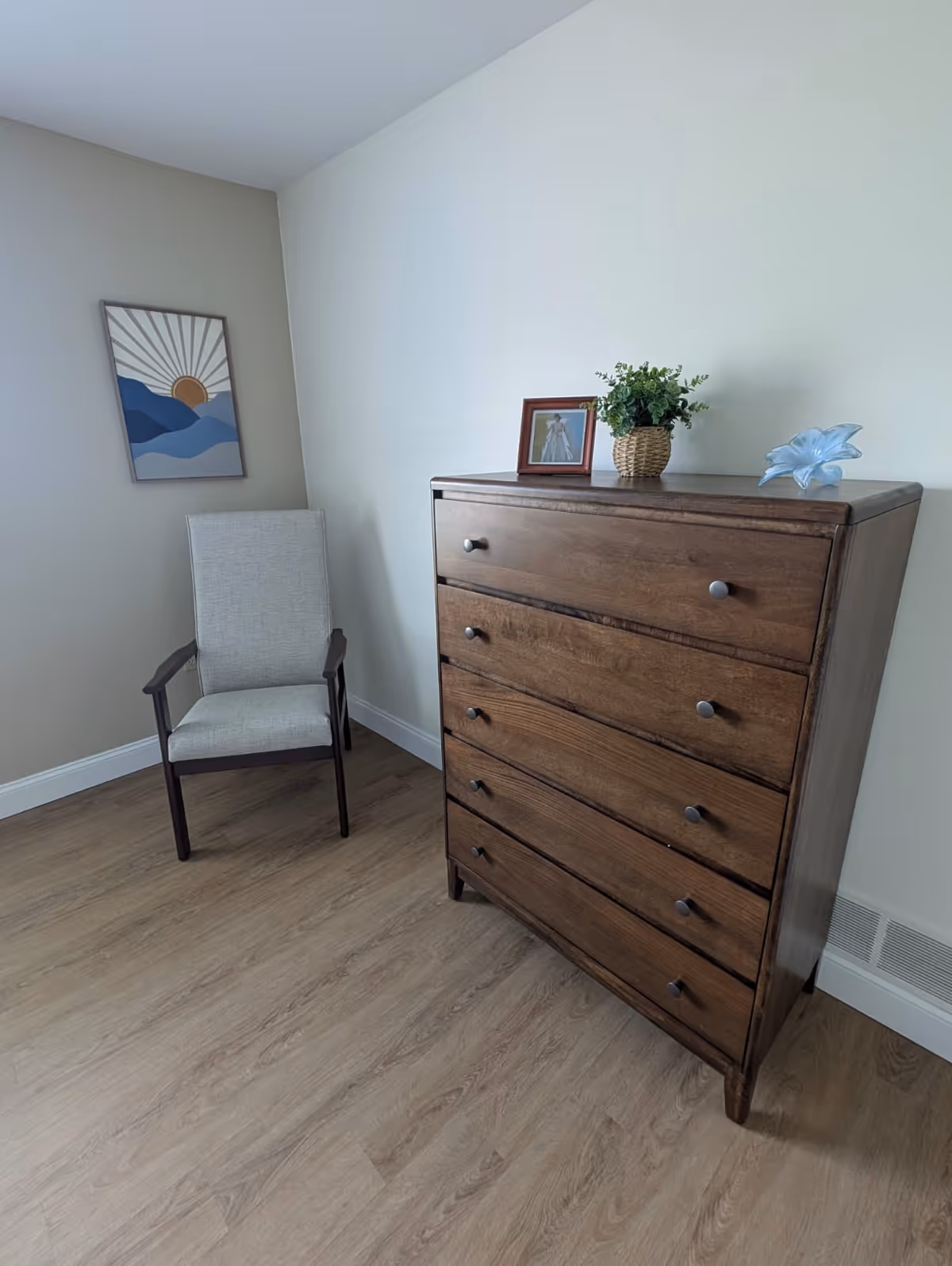A corner of a room with a wooden chest of drawers topped with a small framed photo, a potted plant, and a decorative glass flower. Next to the chest is a light gray upholstered chair with dark wooden arms and legs. A framed abstract artwork featuring a sun and blue mountains hangs on the beige wall above the chair. The floor is light wood, and the walls are painted in light neutral tones.