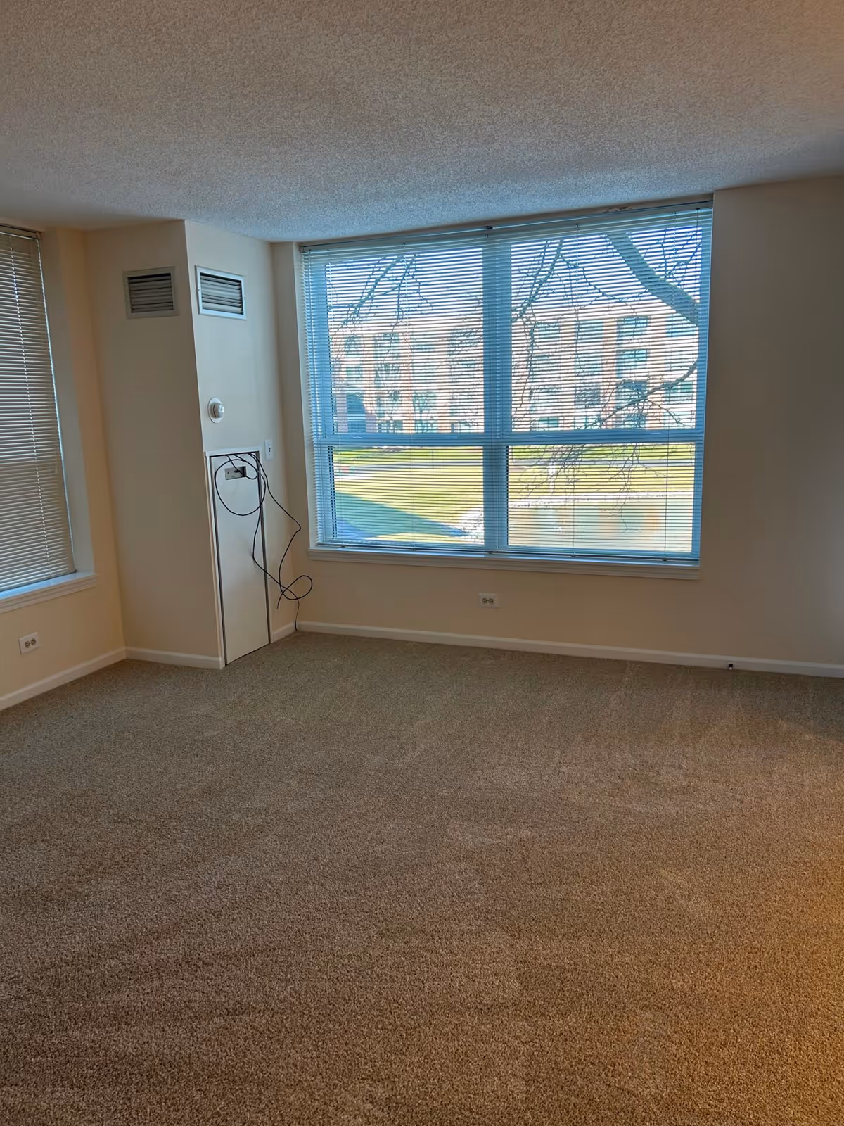 Empty carpeted room with a large window and blinds overlooking a lawn and neighboring building.