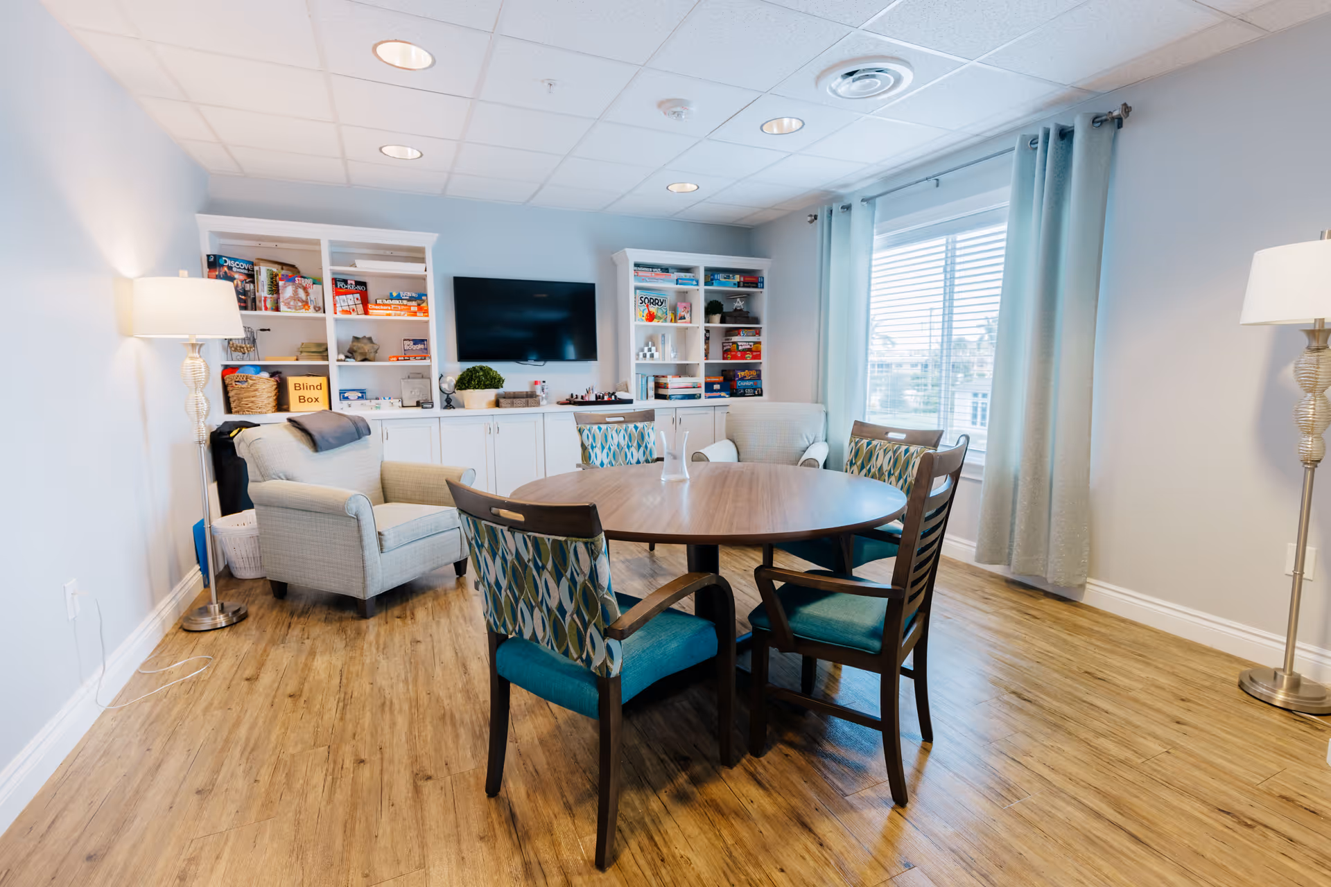 A bright and cozy common room with a round wooden table surrounded by four chairs with patterned cushions. Two armchairs are placed near a white built-in cabinet filled with board games and books. A flat-screen TV is mounted on the wall above the cabinet. The room has wood flooring, light blue walls, two floor lamps, and a large window with light curtains letting in natural light.