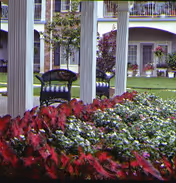 Landscaped courtyard with red and white flowering plants, white columns, a wicker chair, and a multi-story building with balconies in the background.