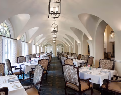 Elegant dining room with rows of tables covered in white tablecloths, set with plates, glasses, and silverware. The room features arched ceilings with hanging lantern-style light fixtures and large windows with patterned curtains allowing natural light to fill the space. Upholstered chairs with intricate designs surround each table.