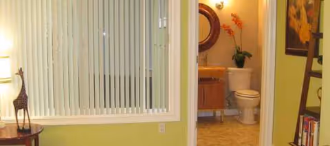 Light green interior with vertical blinds; hallway opens to a bathroom with toilet and sink.