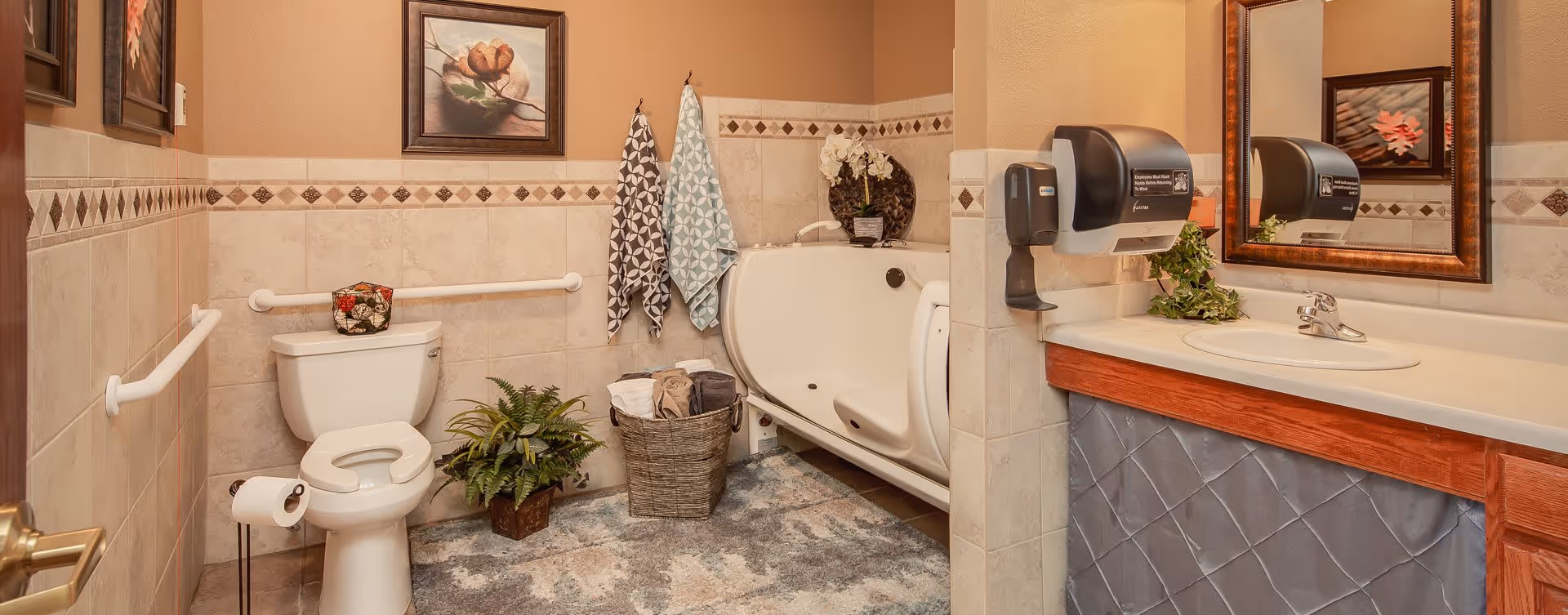 A well-decorated bathroom featuring a toilet with grab bars, a walk-in bathtub with a basket of towels beside it, a sink with a mirror above, and wall-mounted soap and paper towel dispensers. The walls are tiled halfway up with a decorative border, and there are framed floral pictures and hanging towels on the wall. A small plant is placed near the toilet, and the floor has a patterned rug.