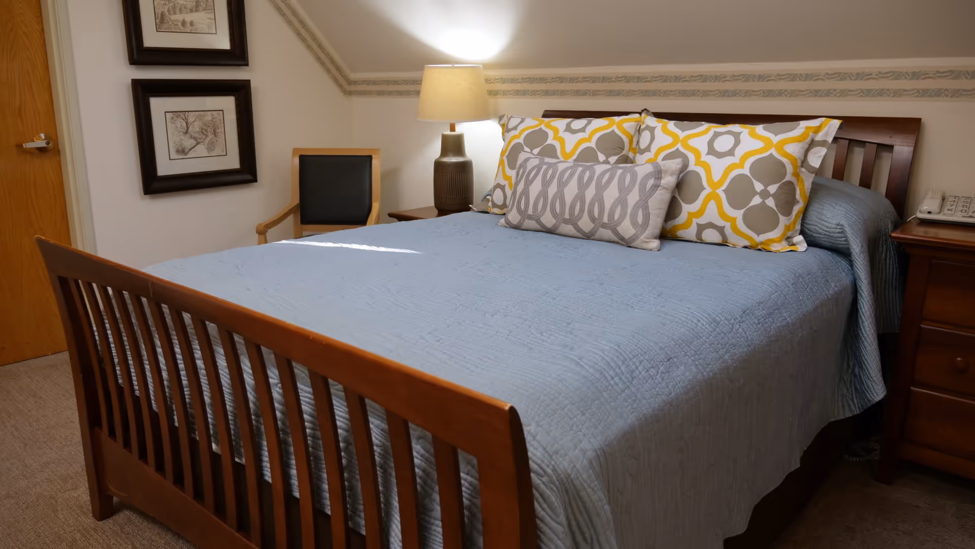 A neatly made bed with a wooden frame, blue quilt, and decorative pillows in a bedroom. There is a wooden nightstand with a lamp and a telephone on the right side of the bed. A wooden chair with a black cushion is placed against the wall, which has two framed pictures hanging above it. The room has beige carpeting and a wooden door on the left side.