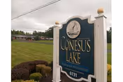 Freestanding decorative sign reading "Conesus Lake" on a grassy lawn with shrubs and the nursing home buildings and overcast sky in the background.