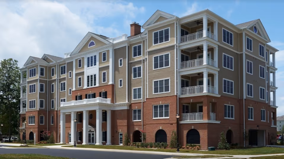 Exterior view of a multi-story senior living facility building with a combination of brick and beige siding, white columns at the entrance, multiple windows, and balconies under a partly cloudy sky.