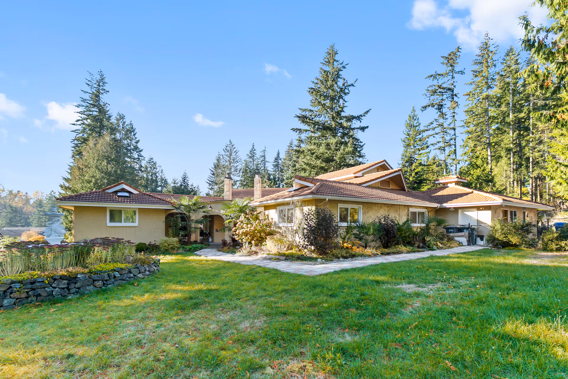 Exterior view of a single-story residential building with a tiled roof surrounded by green grass, landscaped garden beds, and tall pine trees under a clear blue sky.