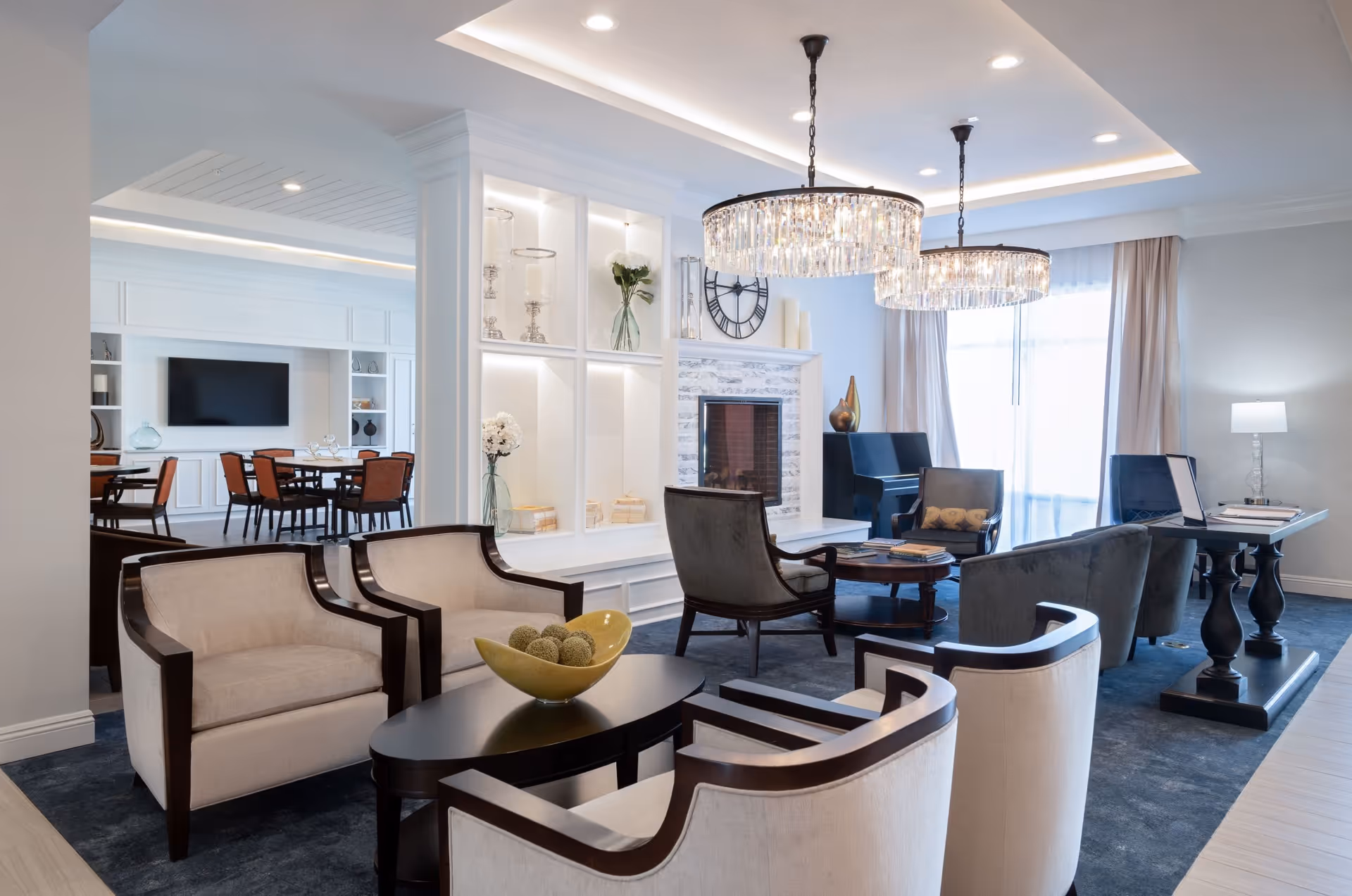 A bright and elegant living room area in a senior living facility featuring beige armchairs with dark wooden frames arranged around a dark oval coffee table with a decorative bowl. In the background, there is a white fireplace with a large clock above it, two crystal chandeliers hanging from the ceiling, a piano, and a table with a lamp near large windows with sheer curtains. Adjacent to the living area is a dining space with a large TV mounted on the wall and several dining tables with chairs.