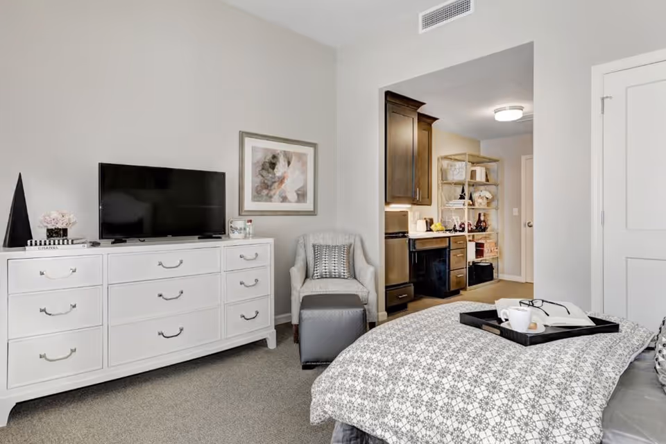 A furnished bedroom with a dresser and TV, armchair, bed with a serving tray, and a small kitchenette visible through a doorway.