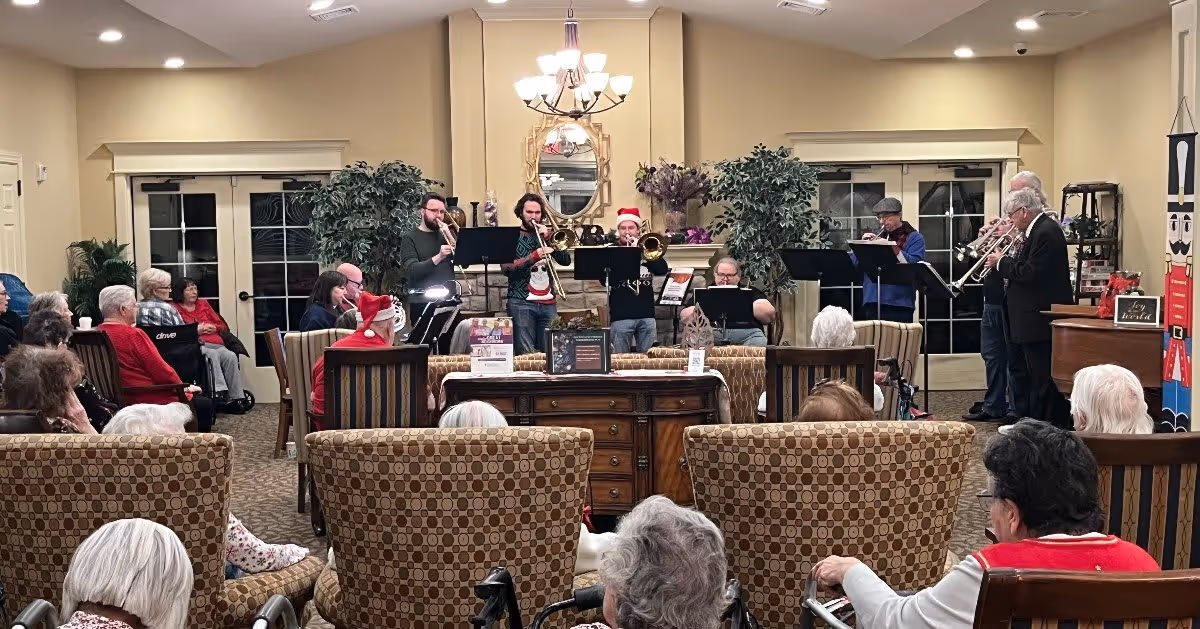 A group of elderly people seated in a living room area watching a small brass band perform. The room is decorated with plants, a mirror above a fireplace, and holiday decorations including a large nutcracker figure. Some audience members are in wheelchairs and a few musicians are wearing festive hats.