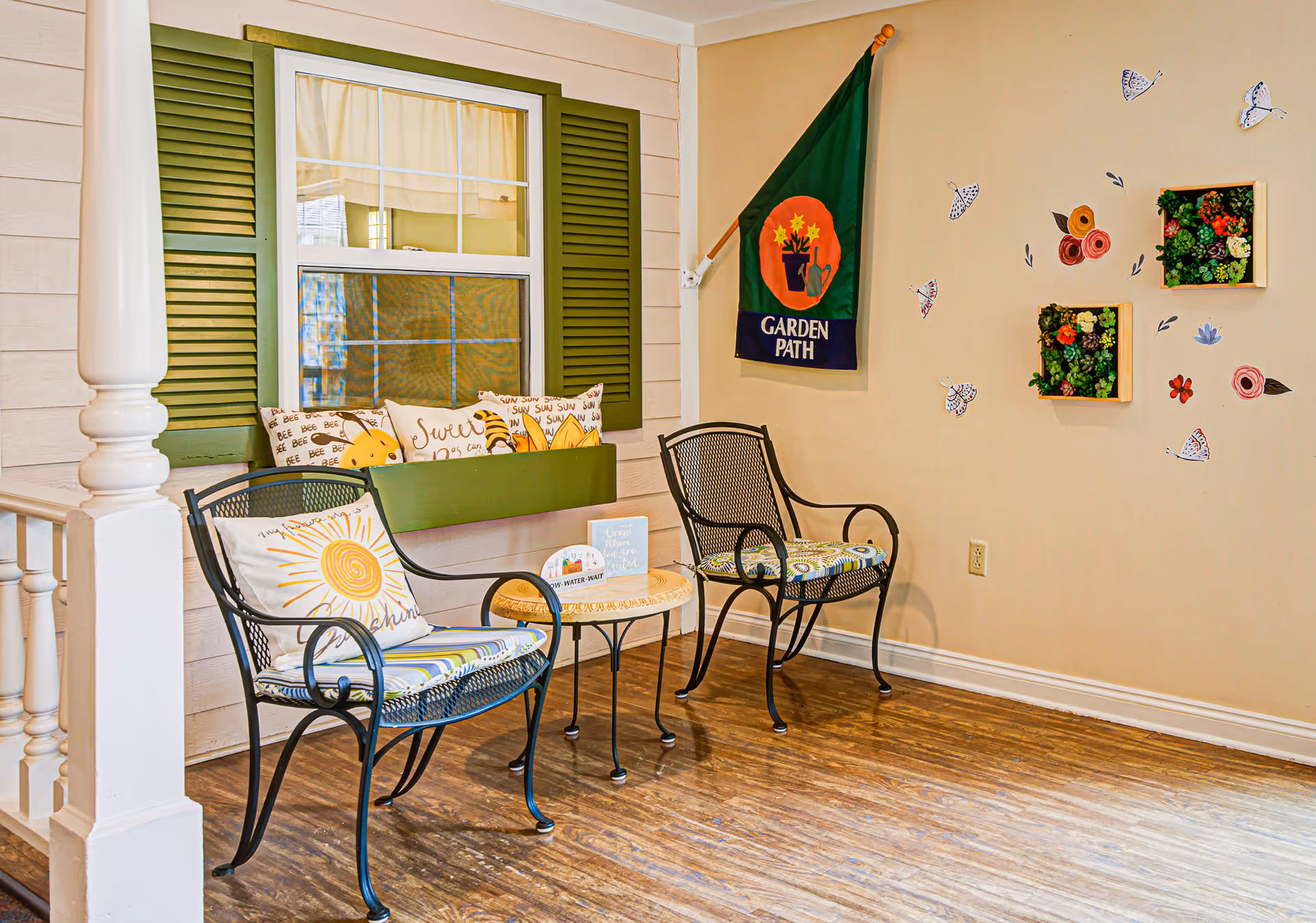 A cozy indoor seating area with two metal chairs featuring patterned cushions and a small round table between them. Behind the chairs is a green bench with decorative pillows, placed in front of a window with green shutters. On the beige wall to the right, there is a flag with the text 'GARDEN PATH' and decorative wall art including small flower boxes and butterfly decals. The floor is wooden, and a white pillar is visible on the left side.