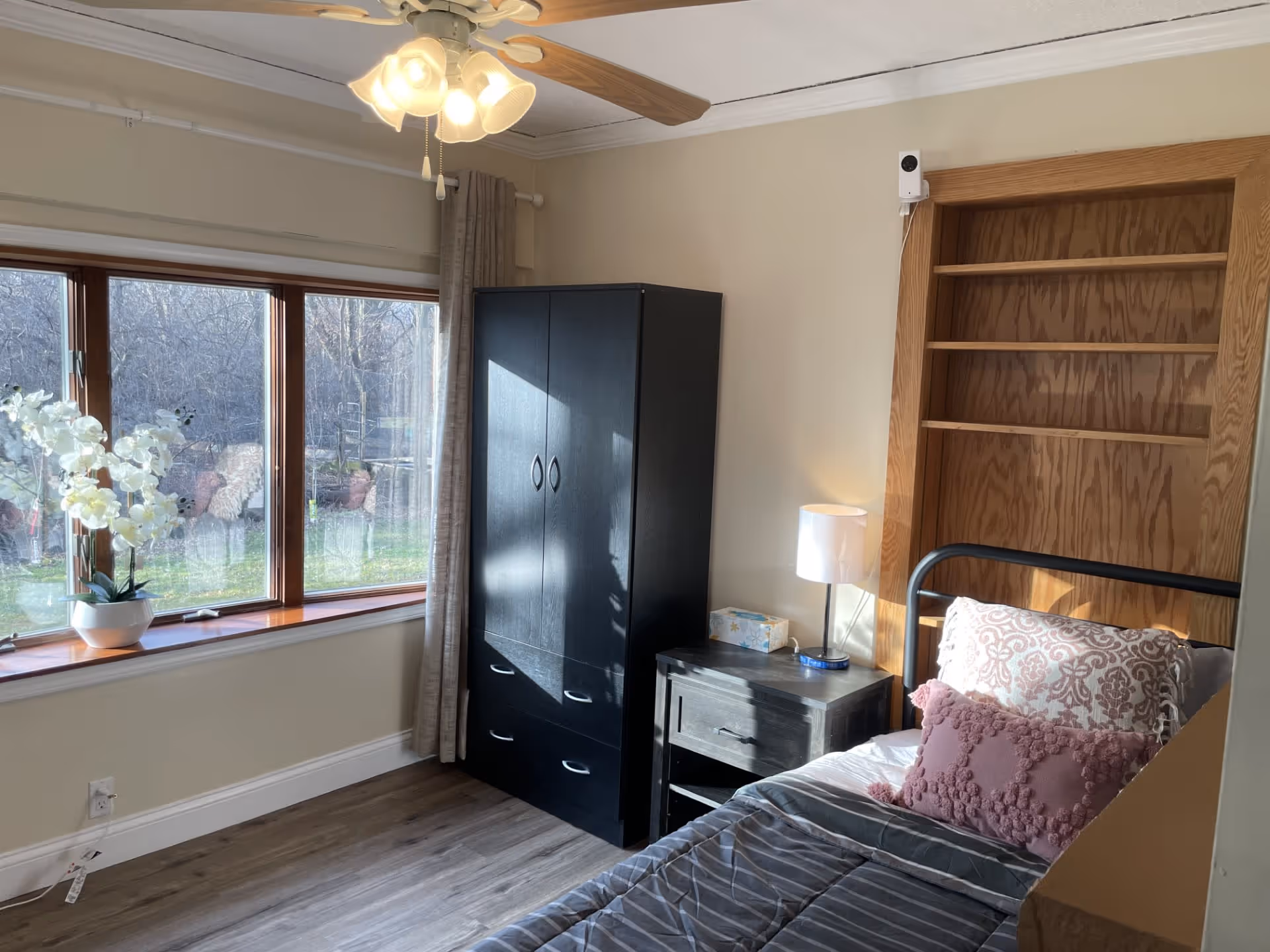 A bright bedroom with a large window letting in natural light. The room features a bed with patterned pillows, a black wardrobe, a small nightstand with a lamp and tissue box, and built-in wooden shelves. A ceiling fan with lights is visible above.