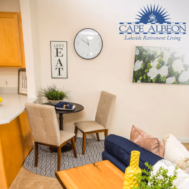 A cozy dining nook with a small round table and two beige upholstered chairs on a patterned rug. The wall behind features a clock, a framed sign that says 'LET'S EAT', and a painting of white flowers. Part of a blue sofa with pillows and a wooden coffee table with a yellow vase and green plant are visible in the foreground. The corner of a kitchen counter with wooden cabinets is also visible.
