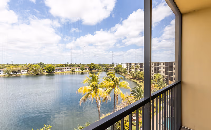 View from a balcony overlooking a lake with palm trees and residential buildings under a partly cloudy sky.
