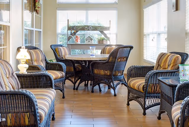 A bright and cozy sitting area with wicker chairs featuring striped cushions arranged around a round table. The room has large windows with blinds allowing natural light to fill the space. A small lamp and decorative items are placed on side tables and a shelf near the window.