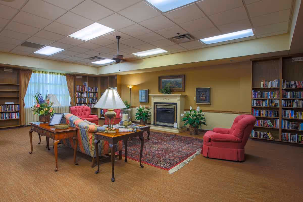 A cozy senior living common area with a floral patterned sofa, two red armchairs, wooden side tables with lamps, a fireplace with framed artwork above, potted plants, bookshelves filled with books, and a large window with curtains letting in natural light.