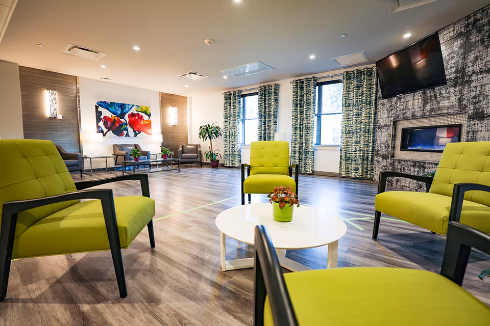 A spacious and modern common area with four bright green armchairs arranged around a white circular coffee table with a small potted plant. The room features wood flooring, large windows with patterned curtains, a wall-mounted flat-screen TV above a contemporary electric fireplace, and colorful abstract artwork on the far wall. Additional seating is visible in the background.