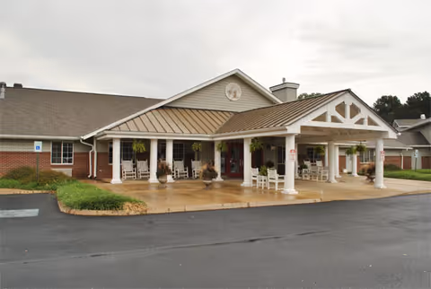 Exterior view of a single-story senior living facility building with a covered entrance supported by white columns. The entrance area has several white rocking chairs and hanging plants. The building has a brick lower facade and beige siding above, with a gabled roof and a circular decorative element near the peak.