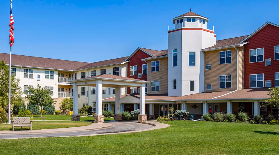 Exterior view of a multi-story senior living facility with a covered entrance, well-maintained lawn, trees, and an American flag on a flagpole against a clear blue sky.