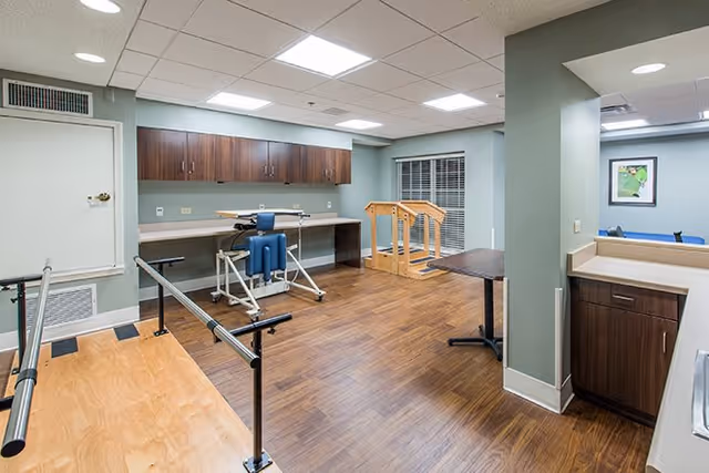 A therapy or rehabilitation room with wooden flooring, parallel bars for walking exercises, a blue therapy chair, wooden handrails, a countertop with cabinets above, and a small sink area. The walls are painted light green and there is a window with blinds.