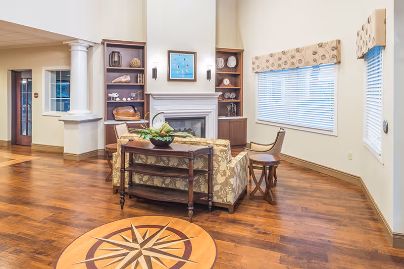 A cozy living room area in an assisted living facility featuring a floral patterned sofa, wooden side table, and a wooden console table with a plant on it. The room has hardwood floors with a compass design inlay, built-in wooden shelves flanking a white fireplace, and large windows with blinds and valances. The walls are painted light beige, and there are wall sconces and a framed artwork above the fireplace.