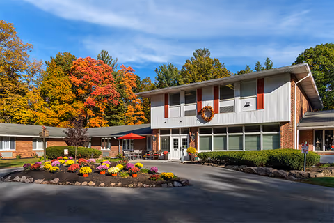 Exterior view of Elderwood Village at Greece facility with a two-story building featuring white siding and red shutters. The entrance is decorated with a fall wreath and surrounded by landscaped flower beds with colorful flowers. Trees with autumn foliage are visible in the background under a blue sky.