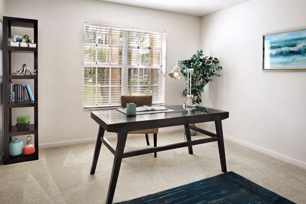 A bright room with a large window covered by white blinds, featuring a dark wooden desk with a glass top, a brown chair, a silver desk lamp, an open book, and a small plant on the desk. To the left is a tall dark wooden shelving unit with decorative items and books. A green leafy plant is in the corner near the desk, and a framed abstract blue and white painting hangs on the right wall. The floor is carpeted with a blue rug partially visible.
