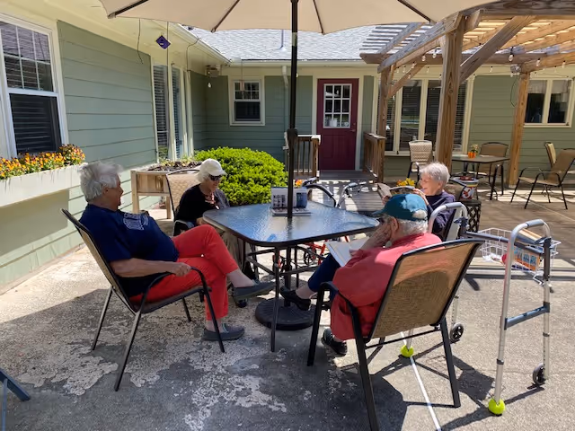 Four elderly individuals sitting around a square outdoor table under a large umbrella in a courtyard area. The setting includes green exterior walls, windows, a red door, and some patio furniture. One person has a walker nearby.