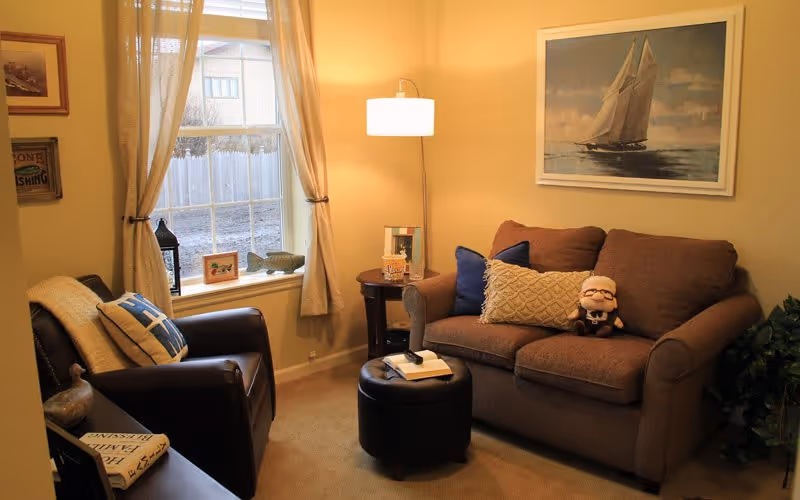 A cozy living room with a brown loveseat adorned with decorative pillows and a small plush doll. Next to it is a round side table with a lamp and a book. Across from the loveseat is a dark brown armchair with a throw blanket and a pillow. A window with beige curtains lets in natural light, and a framed painting of a sailboat hangs on the wall above the loveseat. The room has warm beige walls and carpeted flooring.