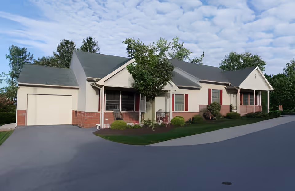Exterior view of a single-story residential building with a garage, green roof, and beige walls with red brick accents. There are several windows with red shutters, a small tree, bushes, and a well-maintained lawn in front. The sky is partly cloudy.