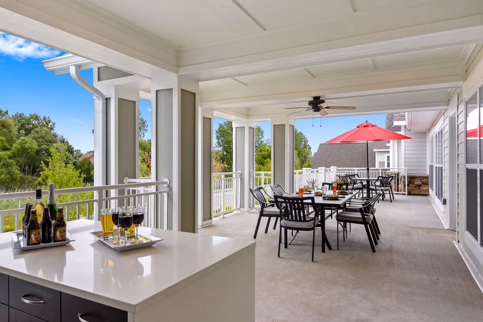 Covered outdoor patio area with tables and chairs, a red umbrella, ceiling fan, and a countertop with trays holding bottles and glasses of beverages. The patio overlooks green trees and a clear blue sky.